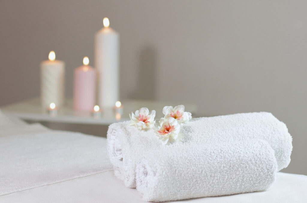 Spa towels are rolled up on top of a massage table. Three blooming flowers sit upon the towels and tall lit candles stand in the background creating a calm and relaxing environment. 