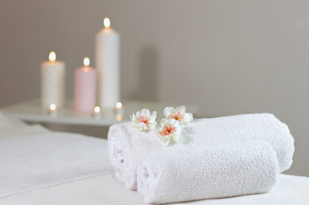 Spa towels are rolled up on top of a massage table. Three blooming flowers sit upon the towels and tall lit candles stand in the background creating a calm and relaxing environment.
