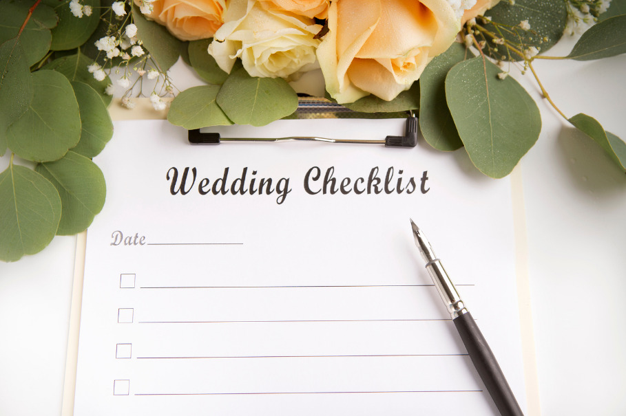 A clipboard holds a piece of paper with “Wedding Checklist” bolded at the top. On top of the clipboard, there are green leaves and pink flowers from a wedding bouquet.