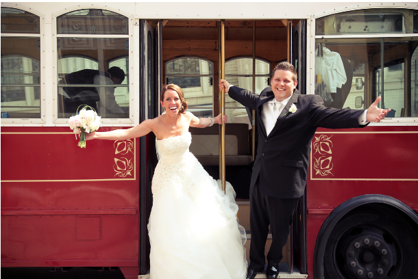 A bride and groom throw up their arms in celebration while on a trolley. 