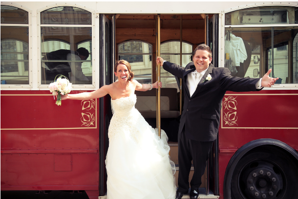 A bride and groom throw up their arms in celebration while on a trolley.