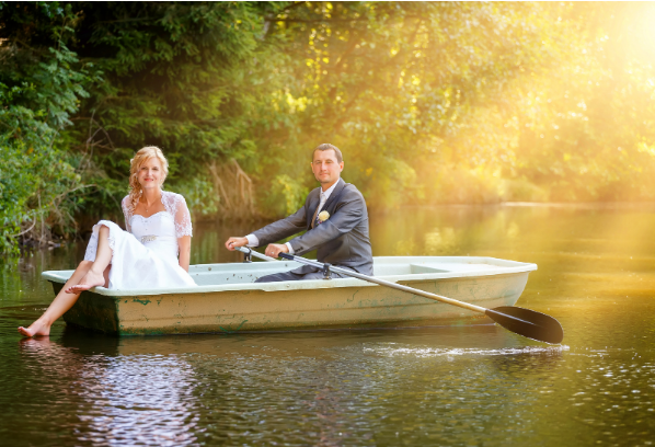 A bride dips her feet in the water while her groom rows the row boat.