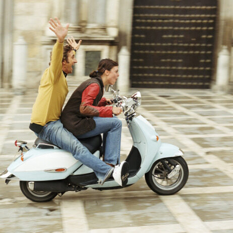 A coupe rides a modep. The woman is driving while the man throws his hands up in excitement. 