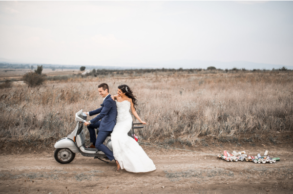 A bride a groom ride a moped. The groom drives as the bride rides behind with her dress waving behind.