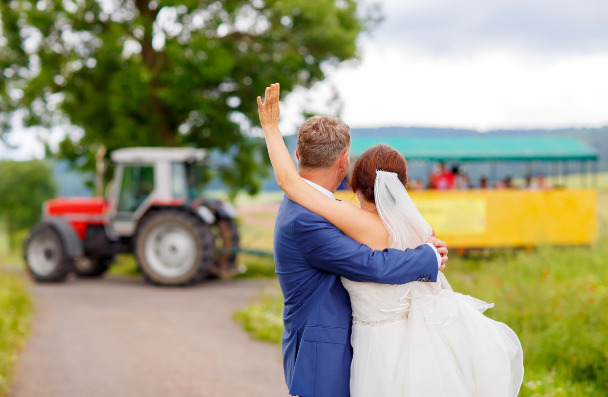 A bride and groom wave to a group of people with a big tractor in the background.