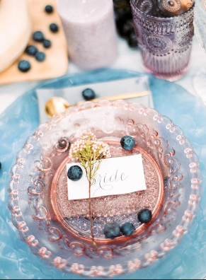 A pastel-colored plate with a nametag resting on top surrounded by berries and a floral stem. The name tag reads “BRIDE”.